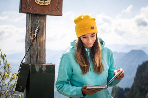 Junge Frau mit gelber Mütze schreibt am Gipfelkreuz in ein Gipfelbuch, im Hintergrund ein Panorama aus Bergen und Himmel.